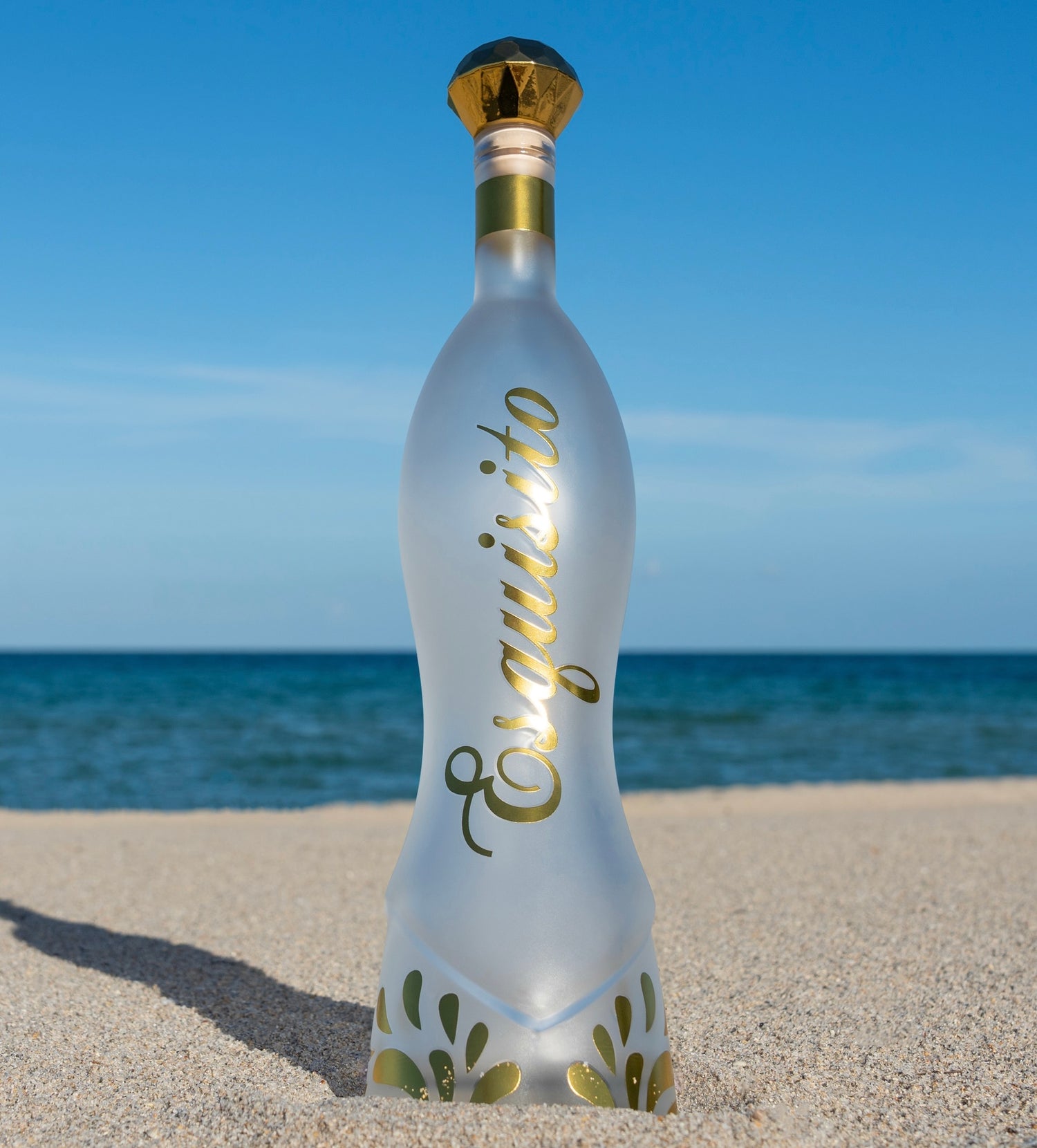 Frosted Esquisto for sipping-bottle with gold accents on a sandy beach with ocean and blue sky in the background